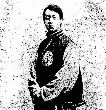 Black-and-white portrait of magician Mac Fee Lung standing with arms folded, wearing traditional-style costume with an embroidered emblem.