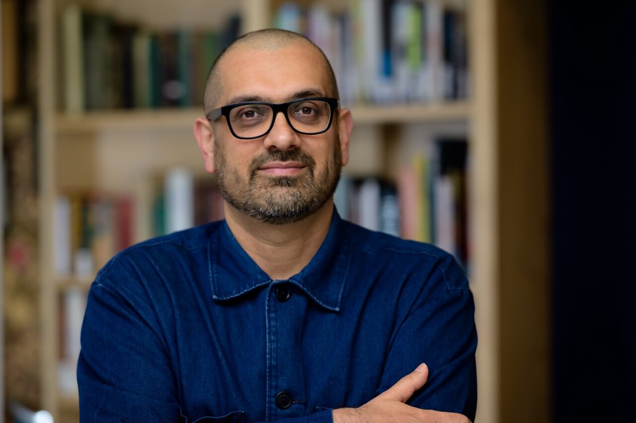 Dr Darren Chetty headshot Portrait of Dr Darren Chetty wearing black glasses and a dark blue shirt, seated with his arms folded in front of softly blurred bookshelves.