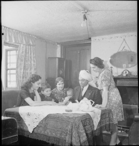 Black and white image of a mixed Muslim family sat around the dining table in the 1940s in a typical British home.