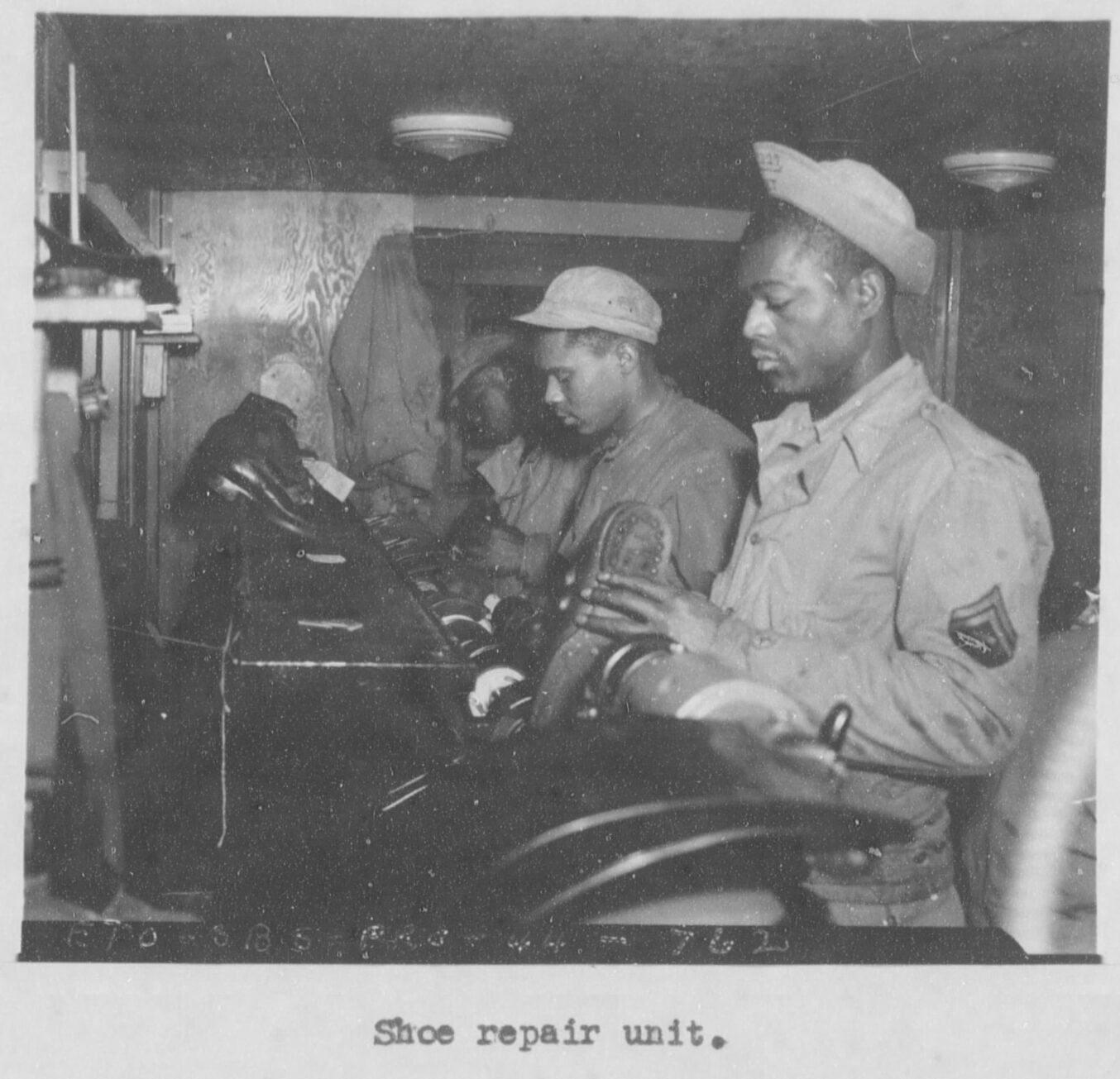 Black and white photo of Black American GIs repairing shoes, Caption reads shoe repair unit.