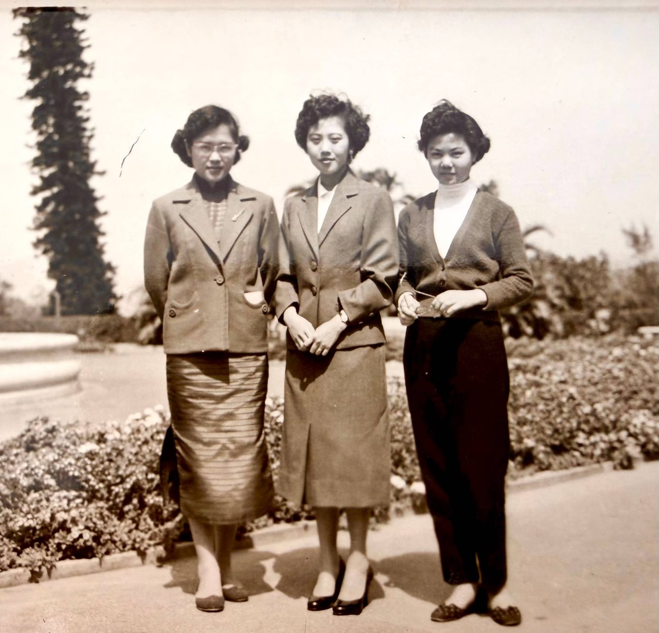 Black and white photograph of Allan O'Neill's mother Yuet Wah posting with two female friends on a road in Hong Kong, 1955. A tree can be seen in the background.