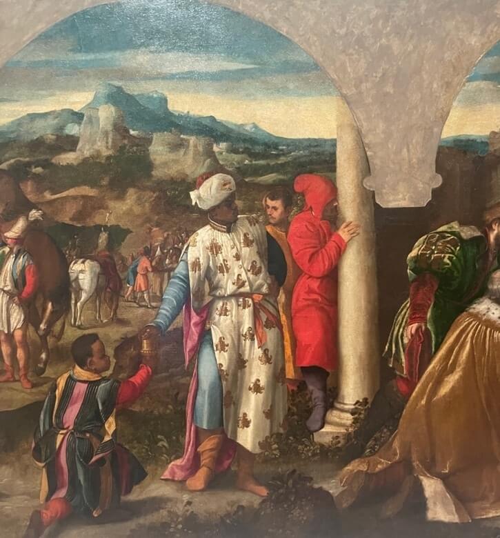 Renaissance painting of the Adoration of the Magi, featuring a Black Magus in ornate dress presenting a gift, surrounded by richly dressed figures with a busy procession and mountainous landscape in the background.