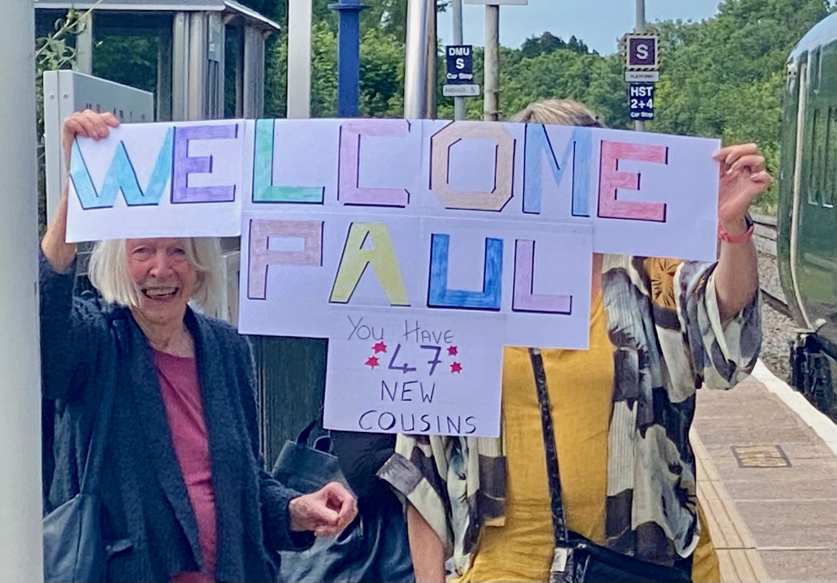 Two women standing on a train platform holding up a sign saying welcome Paul you have 47 new cousins