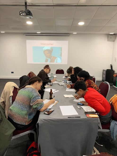 Participants sit around a table writing during an Enter Tragic Mulatta workshop, with a presentation projected on a screen behind them.