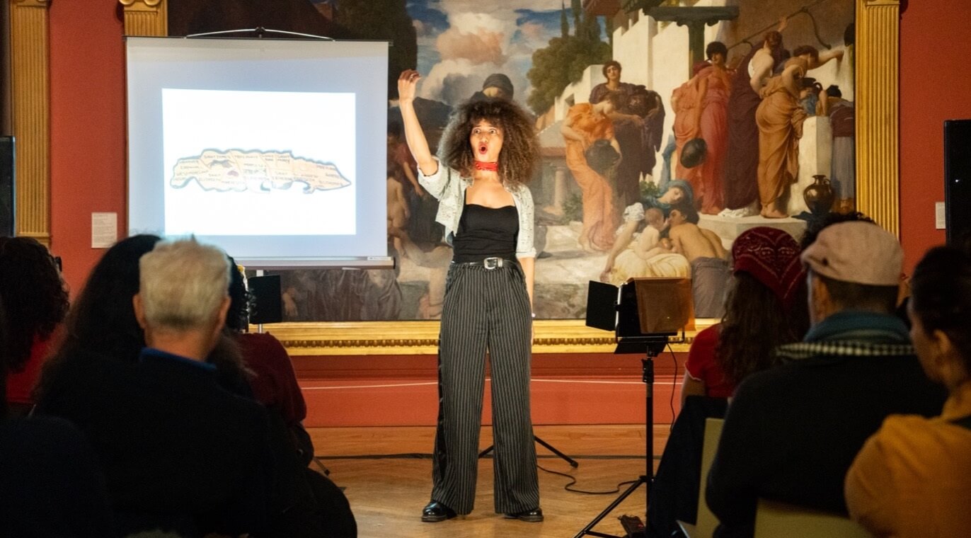 Performer Lauren Fitzpatrick stands before an audience in a gallery, gesturing beside a projected image and large classical painting.