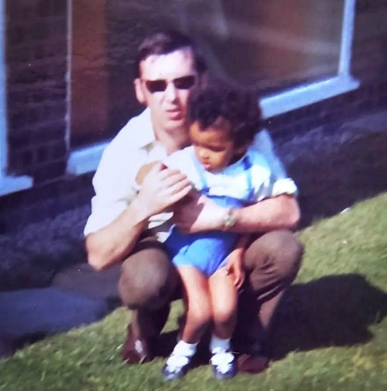 David Yeates as a young boy being hugged by his father who is crouching down. The pair are in a back garden.
