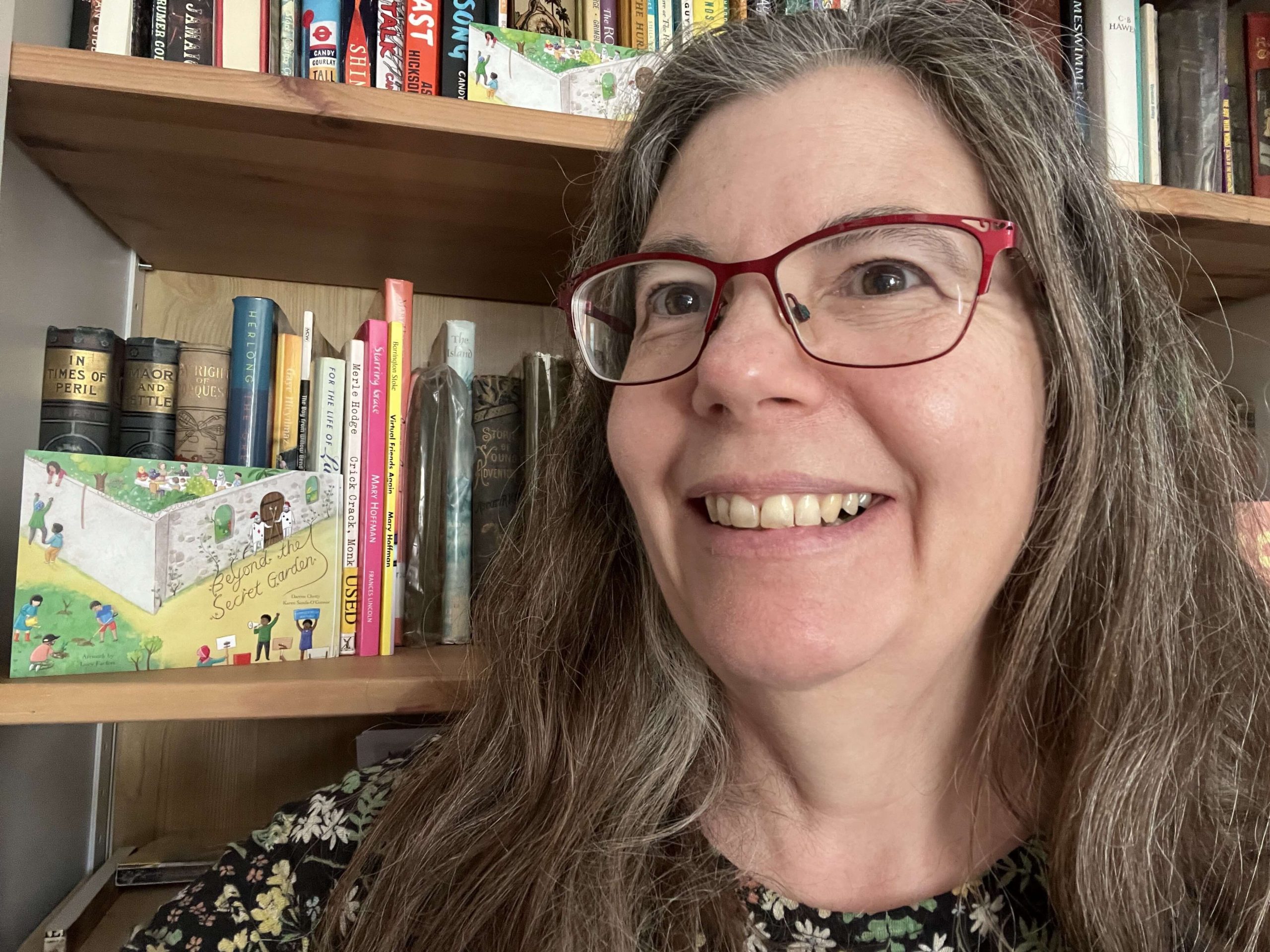 Professor Karen Sands-O'Connor headshot Professor Karen Sands-O’Connor smiling at the camera, wearing red glasses, with bookshelves behind her. A copy of the children’s book Beyond the Secret Garden is displayed on the shelf.