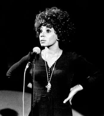 Shirley_Bassey_Rome_1971 Black-and-white photograph of Shirley Bassey performing on stage in 1971, standing at a microphone with one hand on her hip.