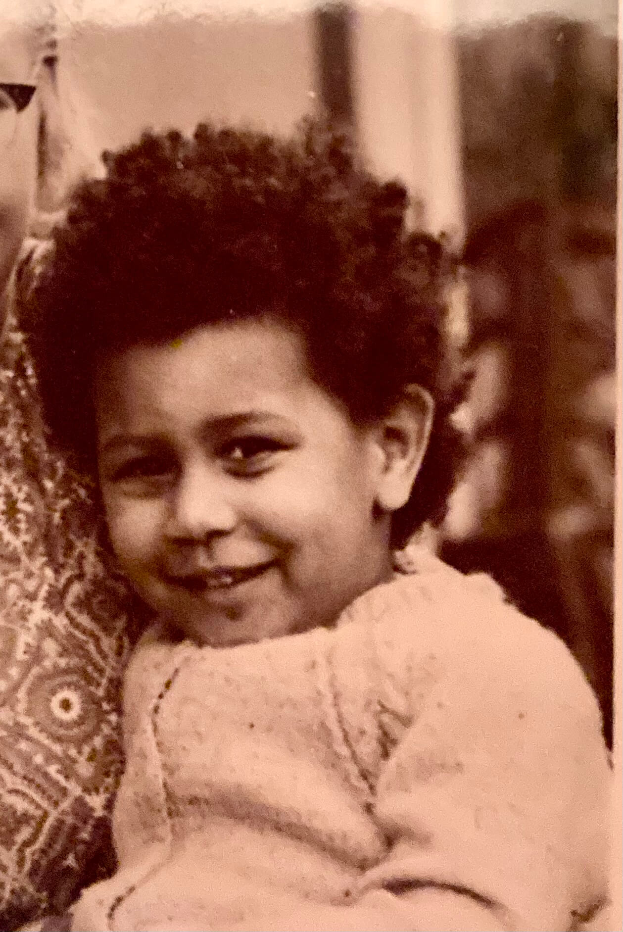 Sepia-toned close-up portrait of Tina Shingler as a young child, smiling while wrapped in a knitted cardigan.