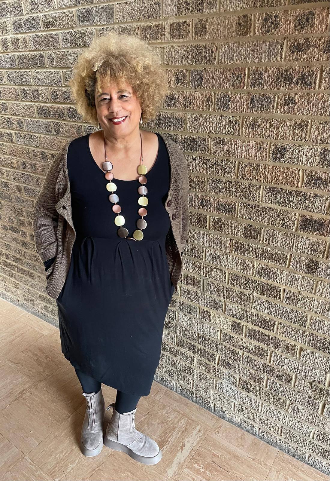 Full-length portrait of Tina Shingler smiling, wearing a black dress, cardigan and long necklace while standing against a brick wall.