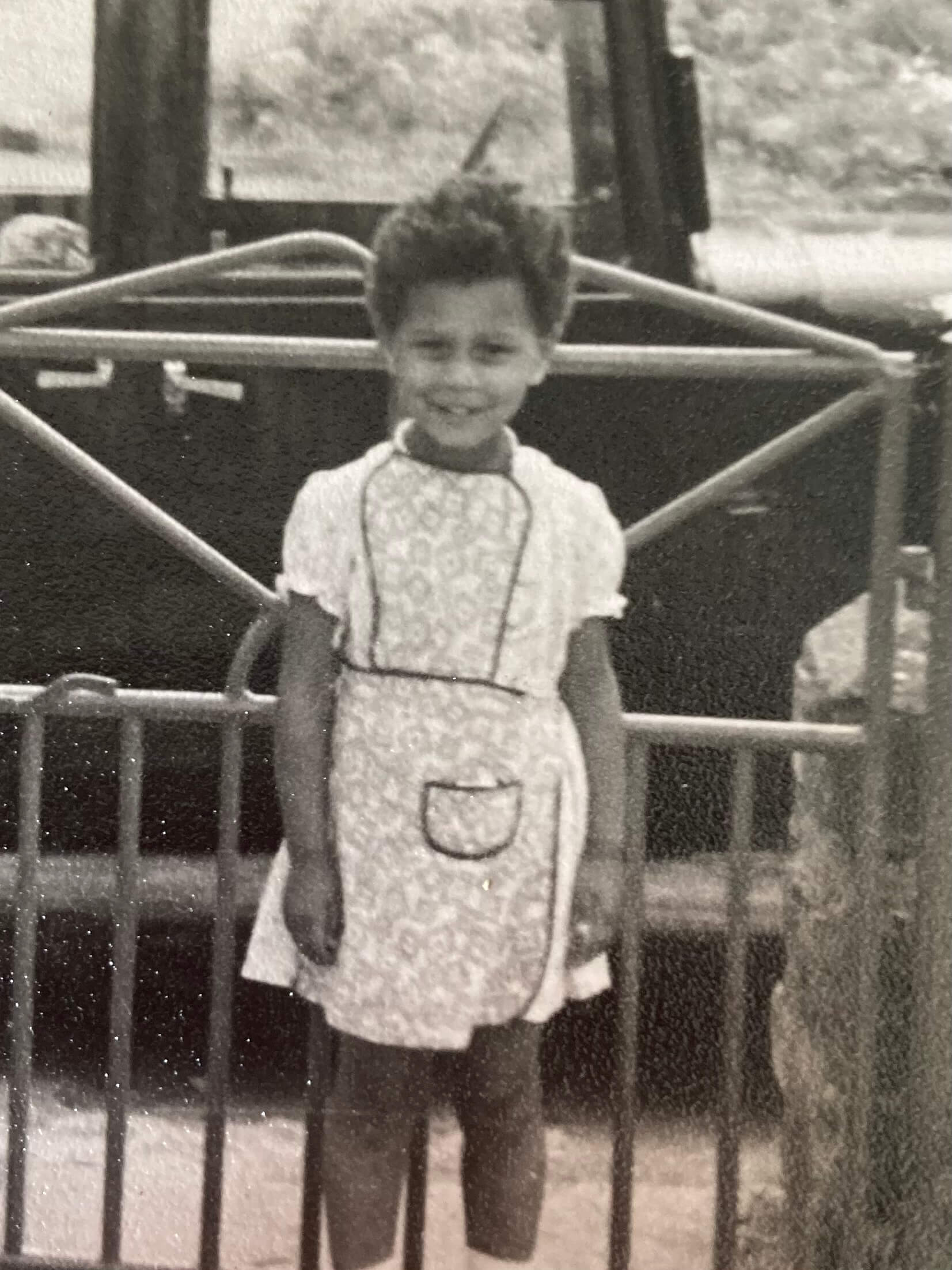 Black-and-white photograph of Tina Shingler as a young child, with short Afro hair, standing by a metal gate in a patterned dress and smiling at the camera.