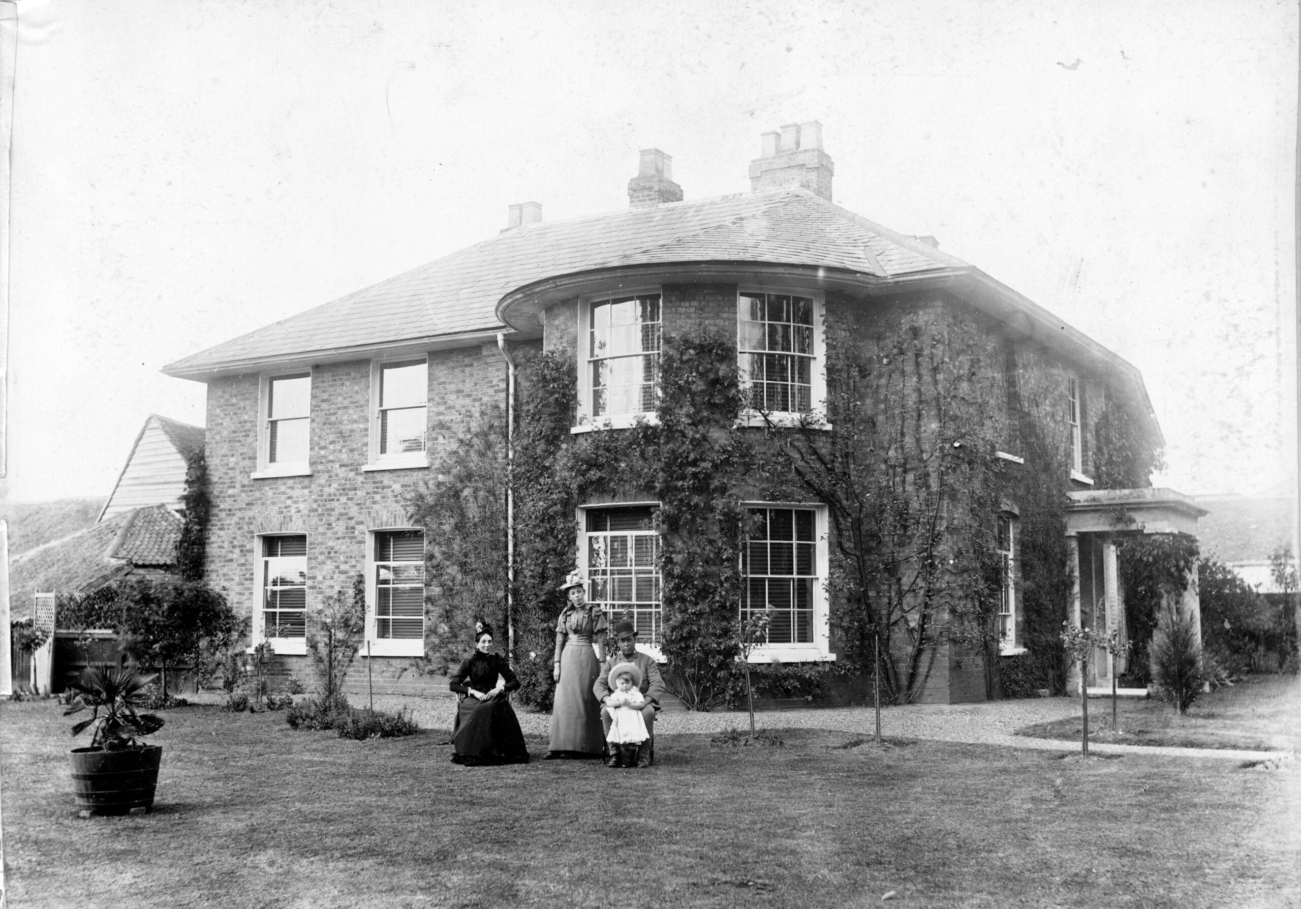 A group of four people posing for the camera on the lawn outside a large house in 1890s Neasden. One woman is seated slightly apart from the other three. The other woman stands next to a man who is seated in a chair with a small child on his lap.