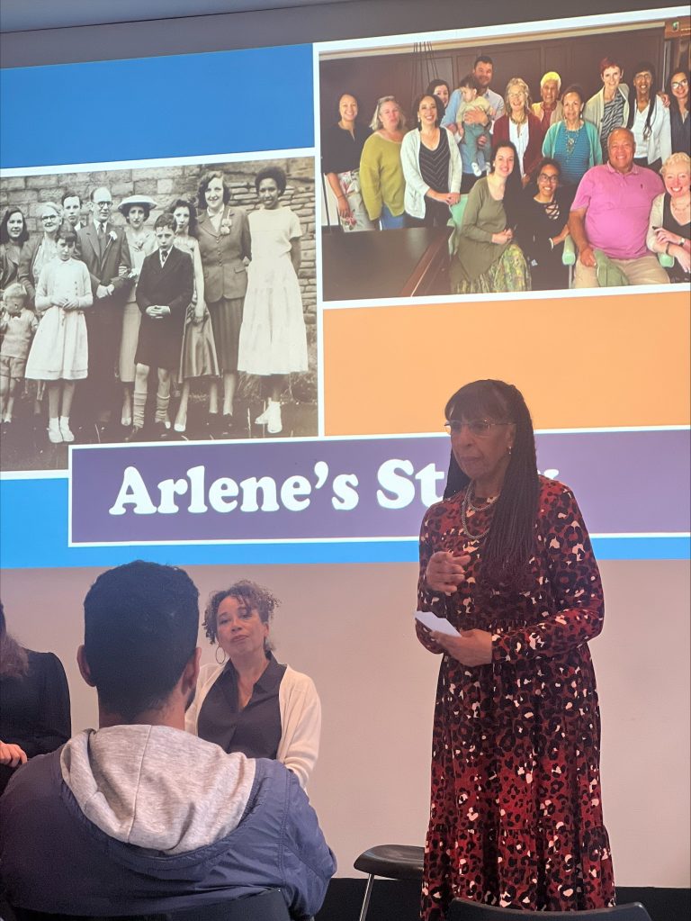 processed-53C6A148-CE31-4A26-9856-E531E9C2AC4F Arlene Nelson talking at the British Science Festival 2025. On the screen behind is a photo of Arlene as a young woman with her family and a photo of Arlene with other 'brown babies' families at The Mixed Museum's Reclaiming History Through Science residential weekend